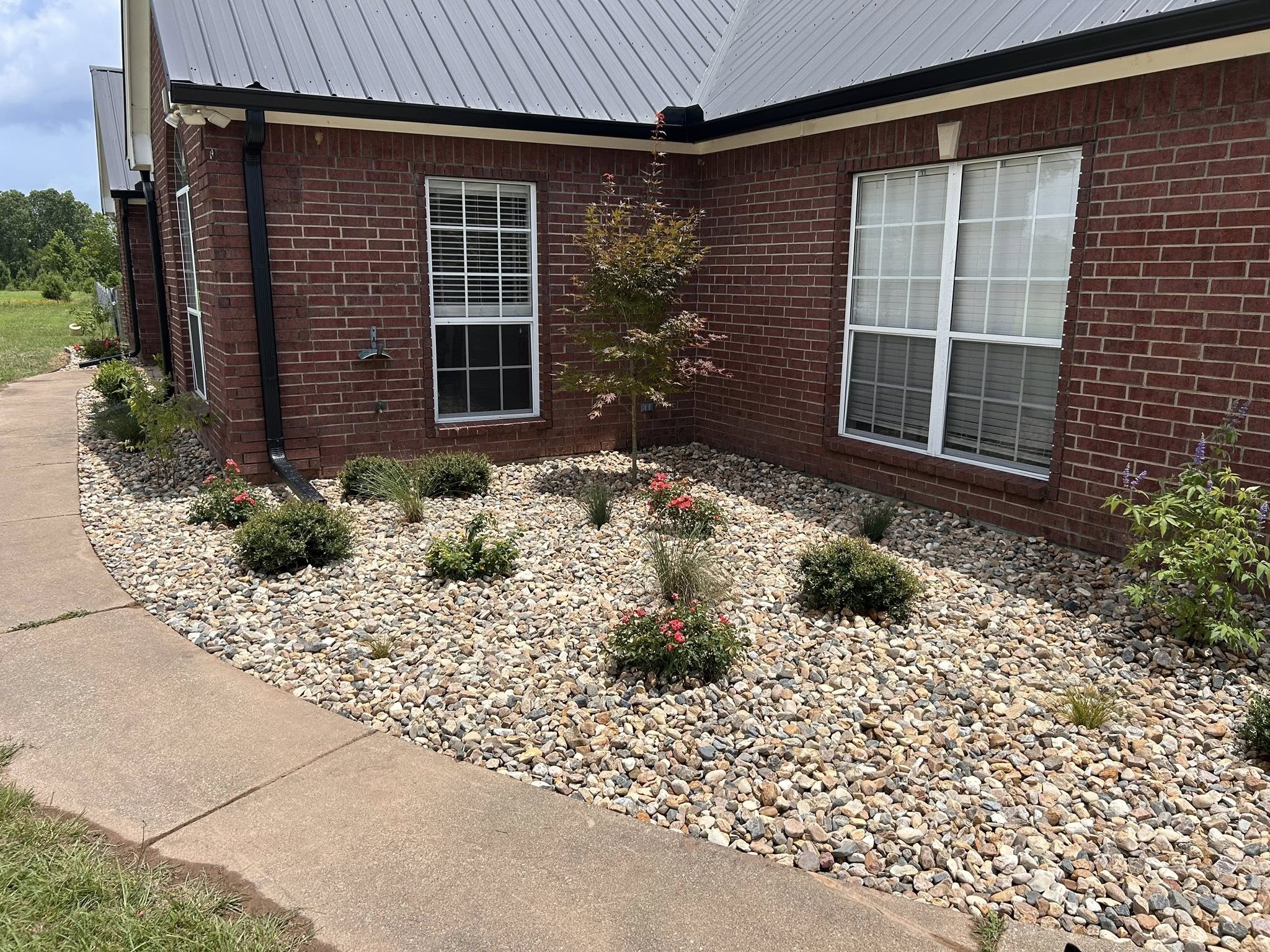Front yard landscaping with decorative river rock, low-maintenance plants, and shrubs along a red brick home – Hood Lawn Care and Services.