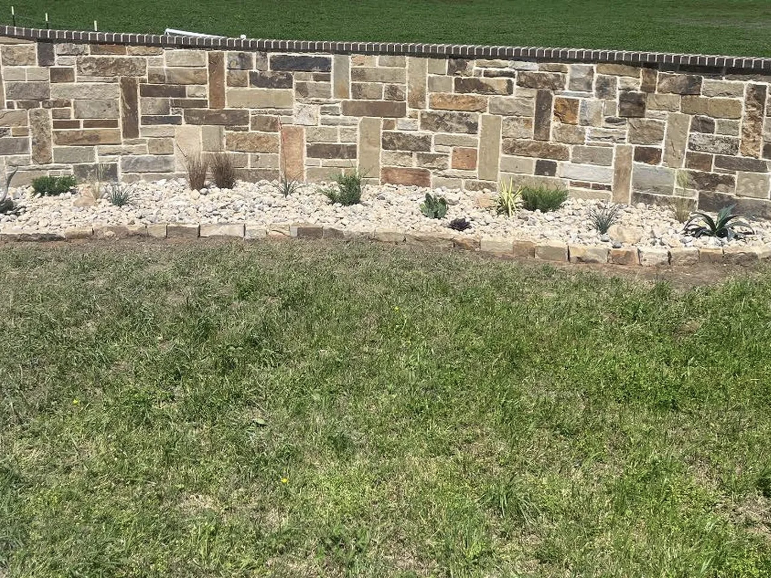 Stone retaining wall with decorative rock flower bed and low-maintenance plants in residential yard – Hood Lawn Care and Services